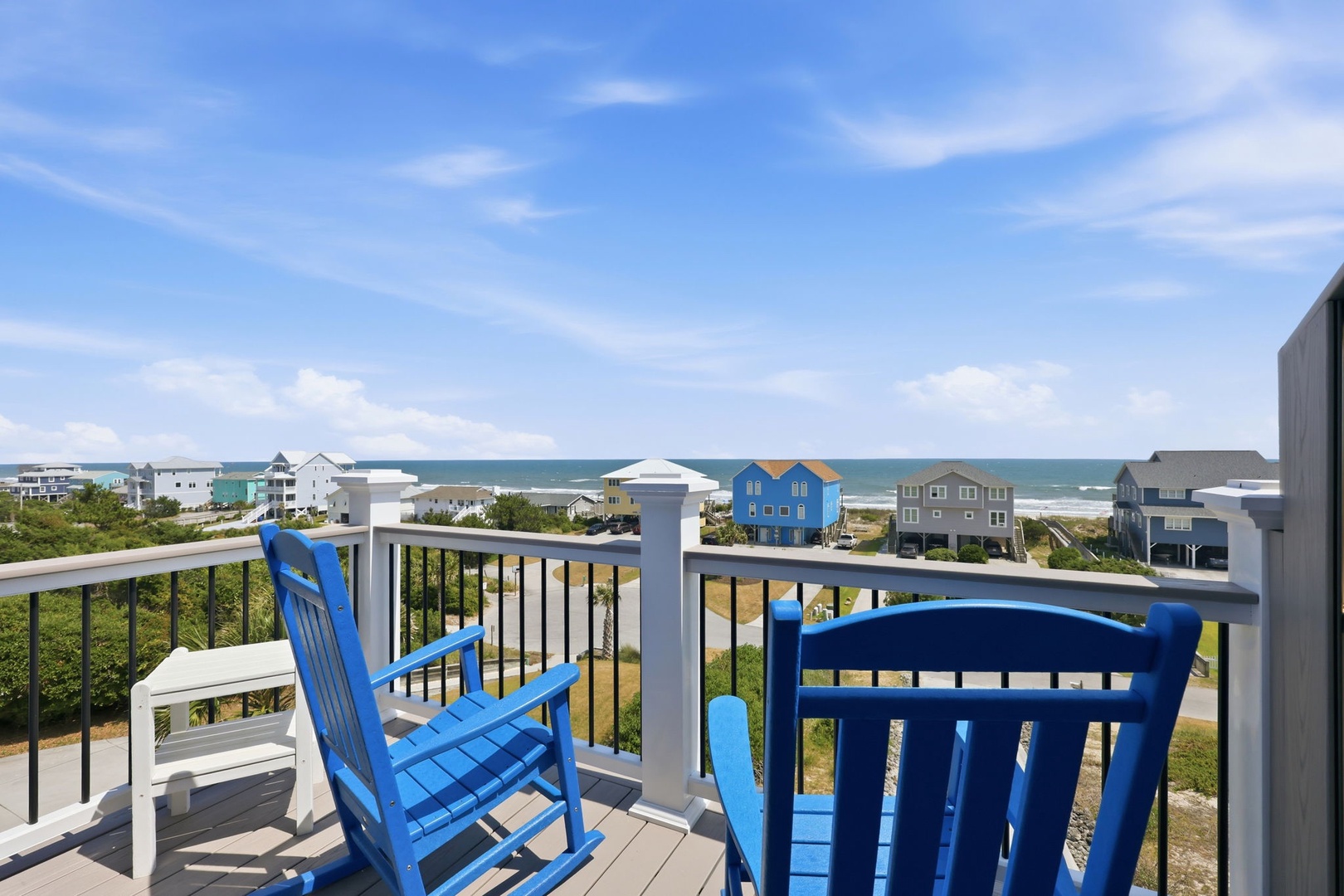 A bright seaside balcony with coastal homes and ocean views stretching to the horizon, framed by blue skies and lush vegetation.