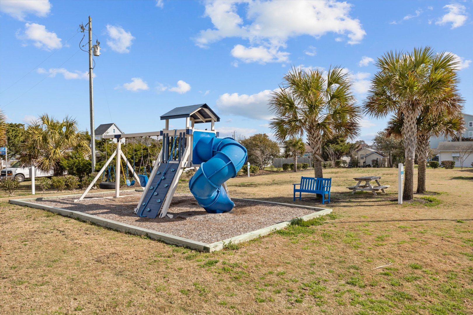 Community playground features slides, swings, and picnic areas surrounded by palm trees and nearby residential homes.