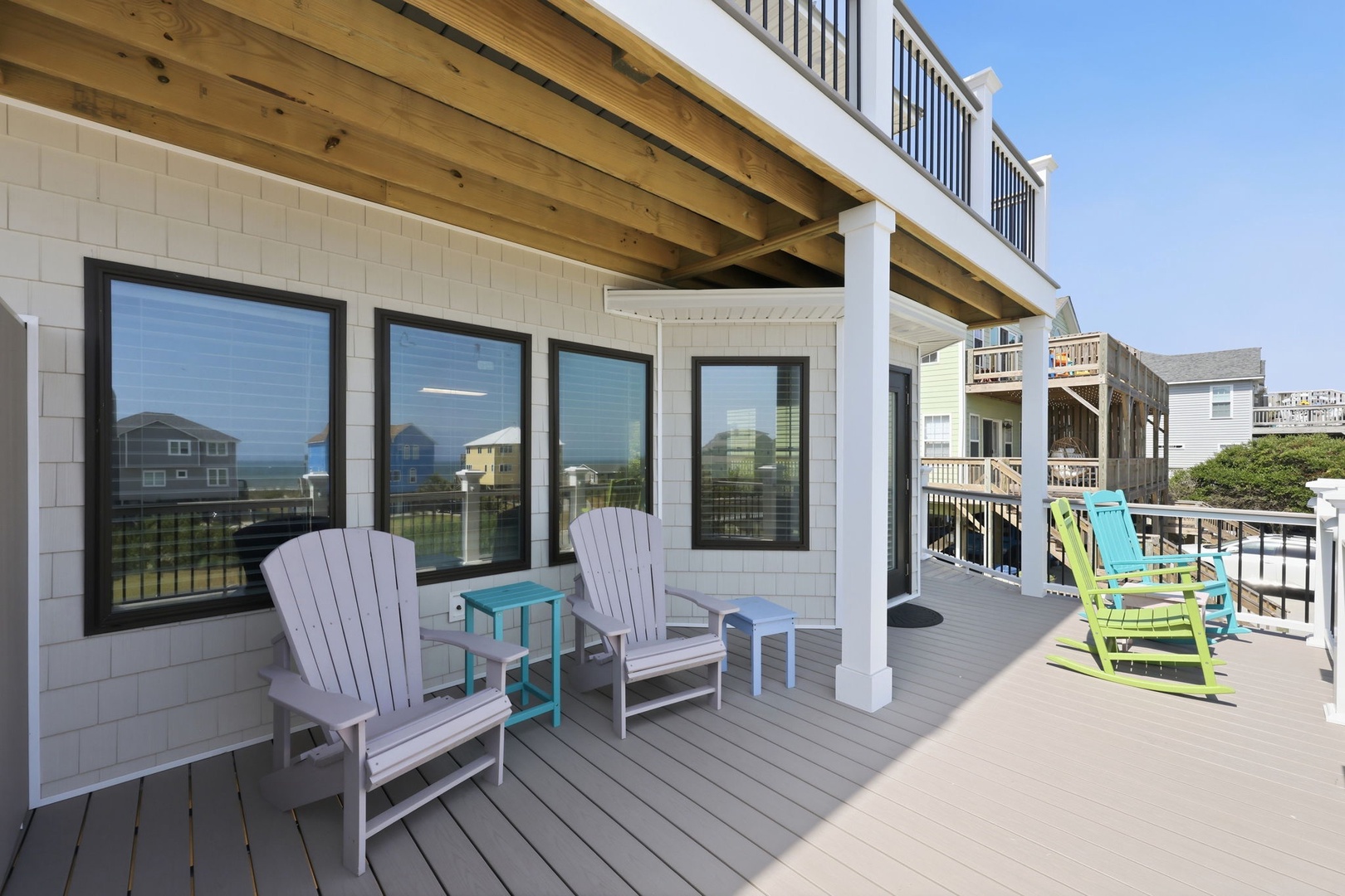 A spacious beach property deck features Adirondack chairs and sun loungers in coastal colors, offering views of surrounding beach houses and natural landscape from this vacation retreat.