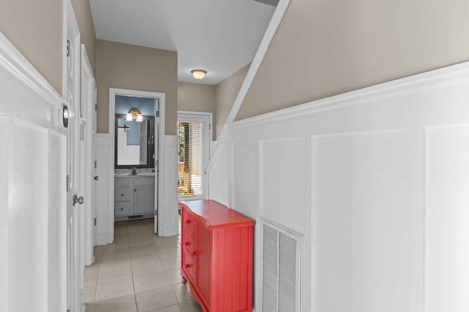 Your charming hallway features elegant white wainscoting and a vibrant red dresser, creating a welcoming coastal atmosphere as you move between spaces.