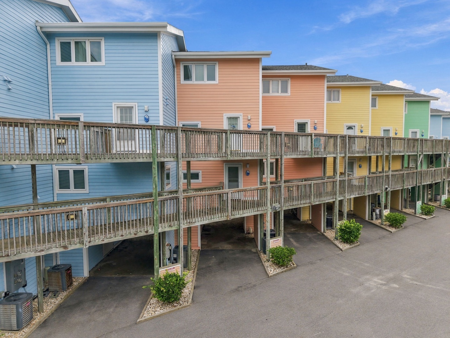 A row of cheerful, brightly-colored beach houses line the property, offering coastal charm with elevated parking and private balconies under sunny skies.