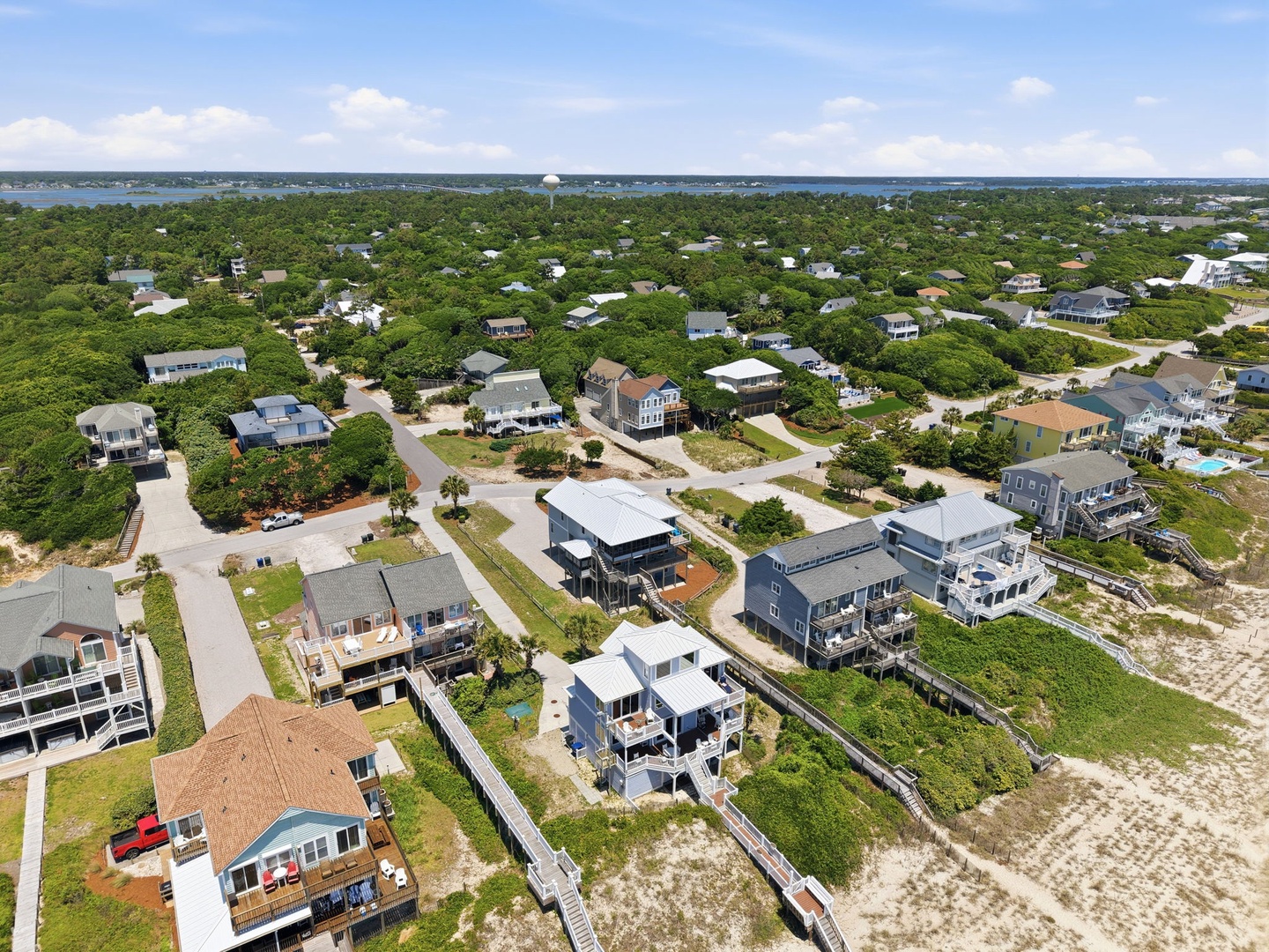 Aerial view of a coastal residential neighborhood featuring vacation homes nestled among lush greenery, with waterfront access and sandy dunes leading to the beach.