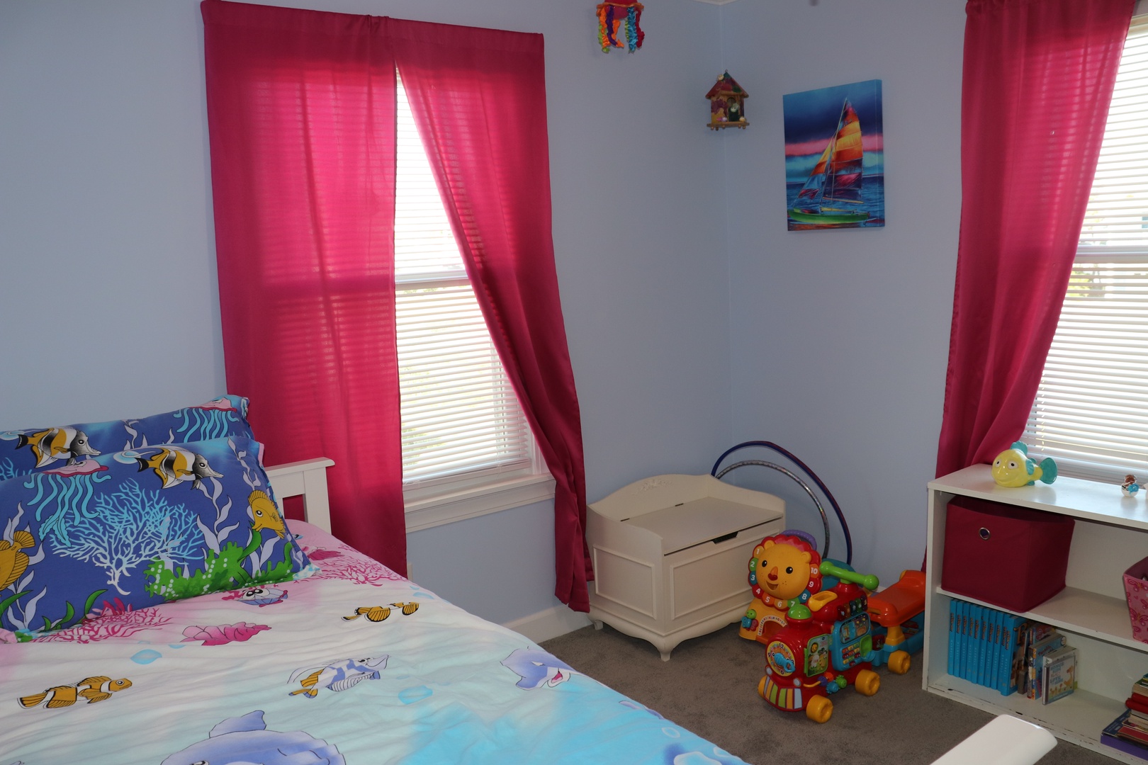 Your little ones will love this cheerful bedroom with ocean-themed bedding, colorful toys, and bright curtains that frame natural light beautifully.