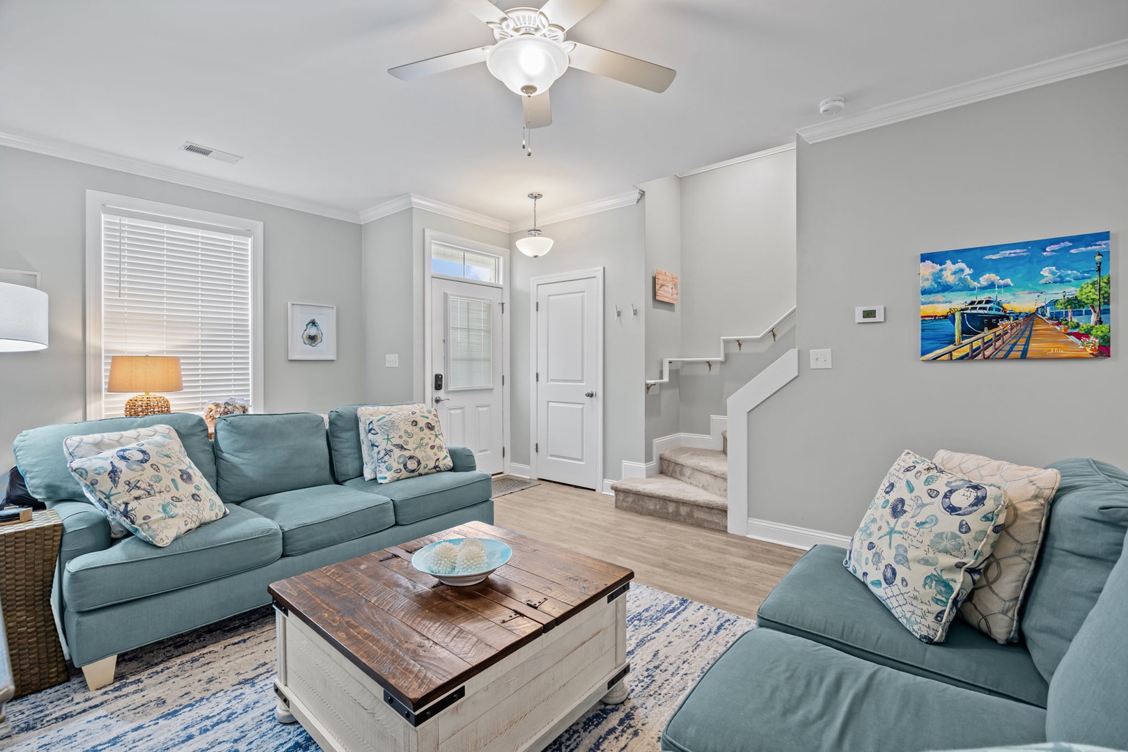 Relax in your bright living room with cozy teal sofas, rustic coffee table, and coastal artwork that sets the perfect beach vacation mood.