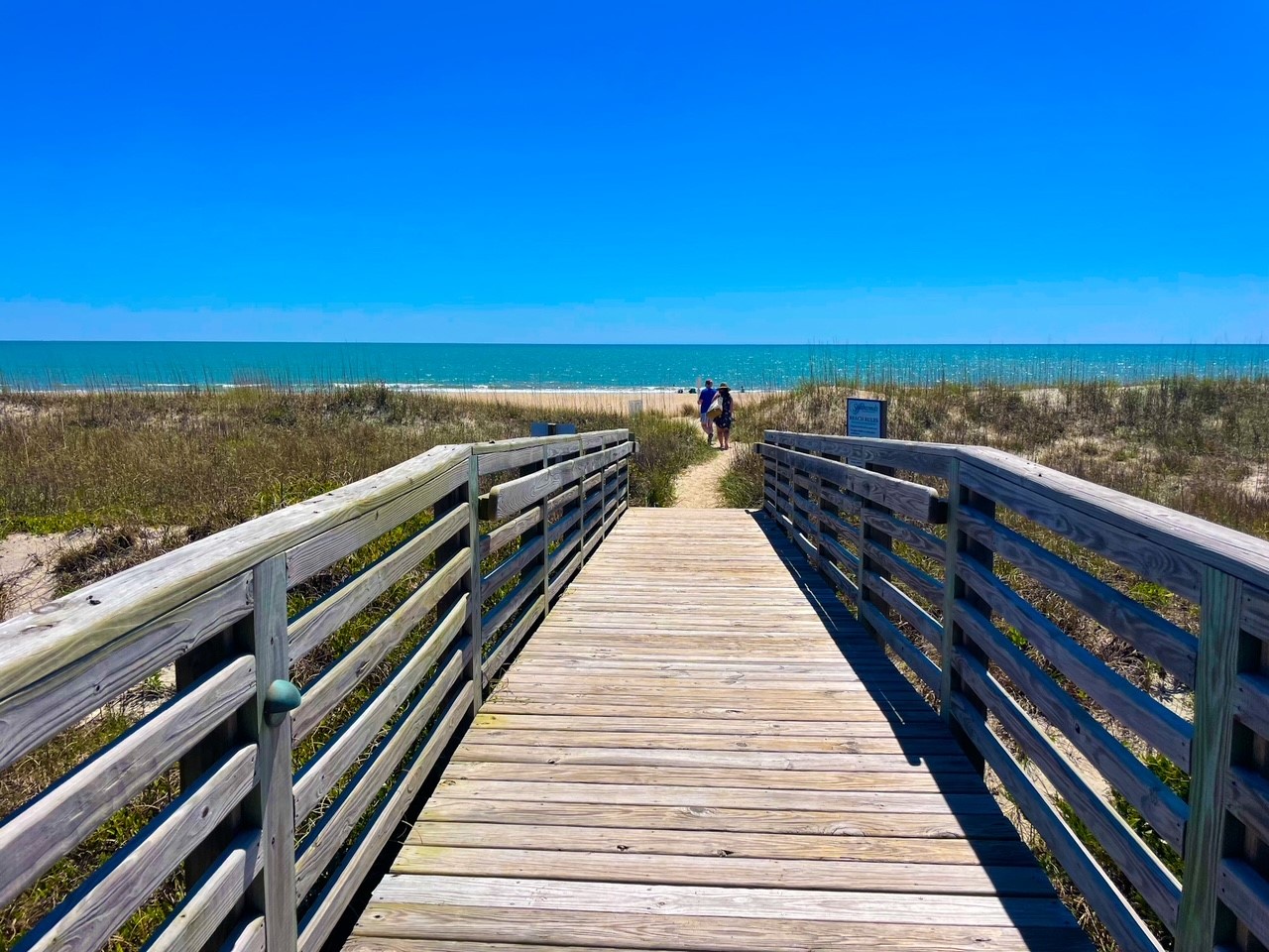 A wooden boardwalk leads through coastal dunes to pristine sandy beach and sparkling ocean waters under brilliant blue skies.