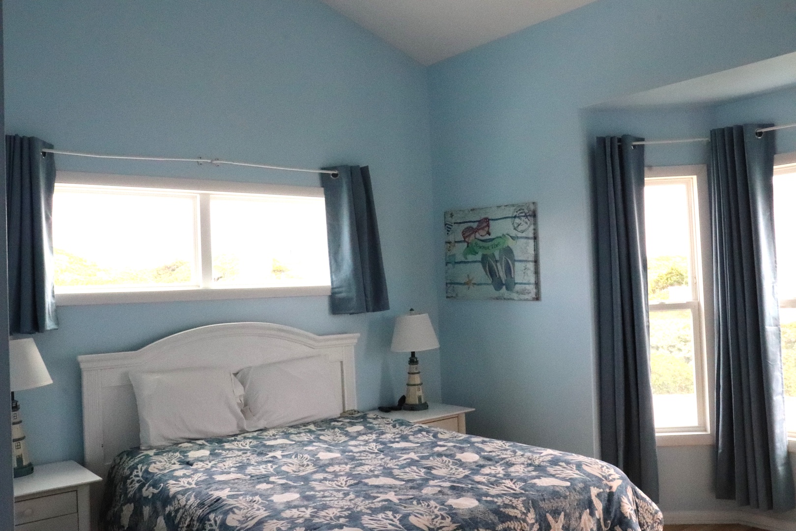 Step into your coastal-inspired bedroom with soothing blue walls and soft natural light streaming through multiple windows. Your comfortable bed awaits beneath charming nautical artwork, creating a serene seaside retreat.