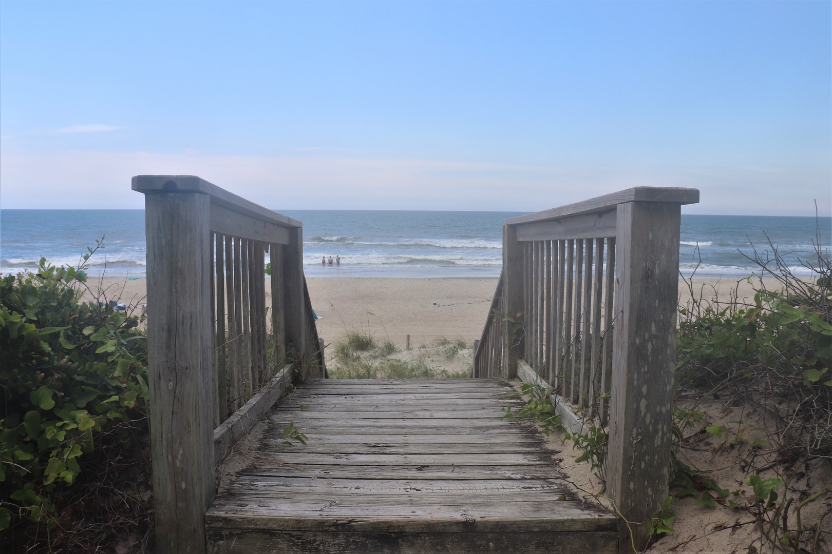Wooden boardwalk leads through coastal vegetation to a pristine sandy beach with gentle waves and clear blue skies.