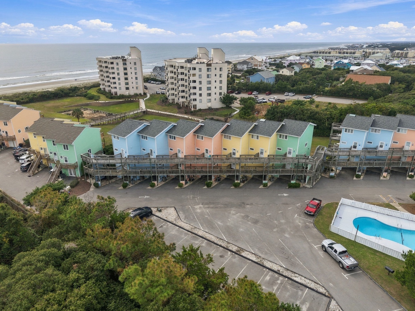 An aerial view of a colorful row of beachfront townhomes near a sandy shore, with a swimming pool and parking area adjacent to the property, situated within a coastal neighborhood.
