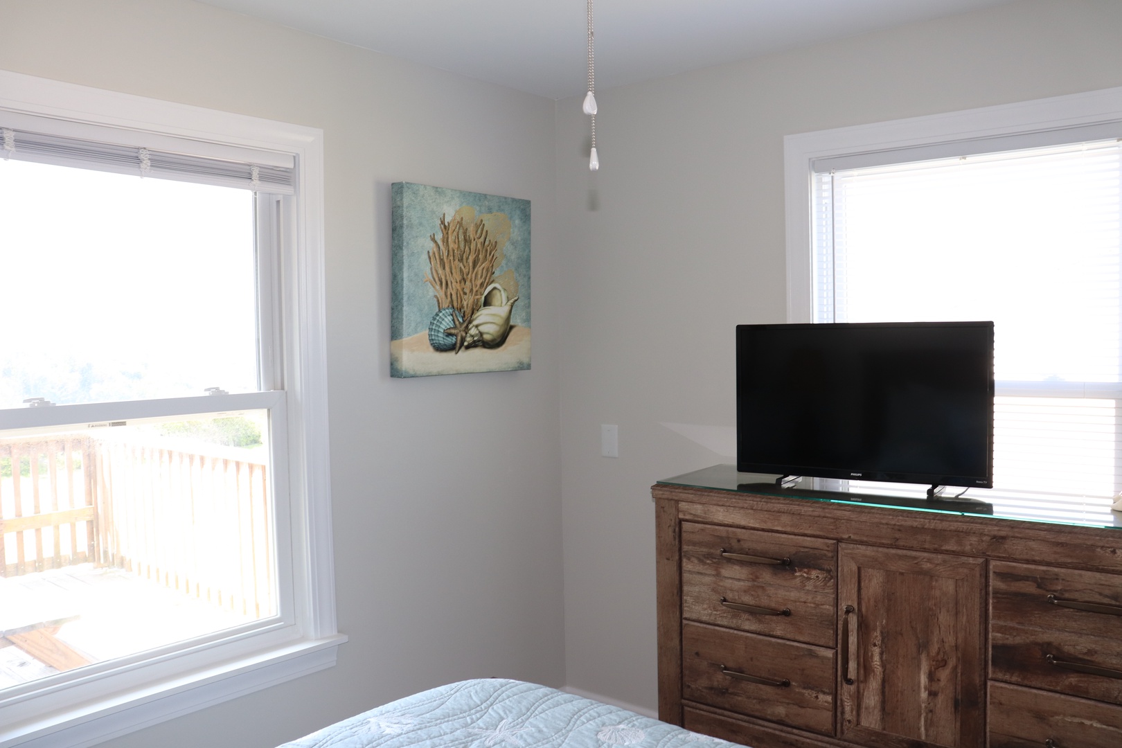 Relax in your bright corner bedroom with natural light streaming through dual windows, featuring a large TV and rustic dresser for your comfort.