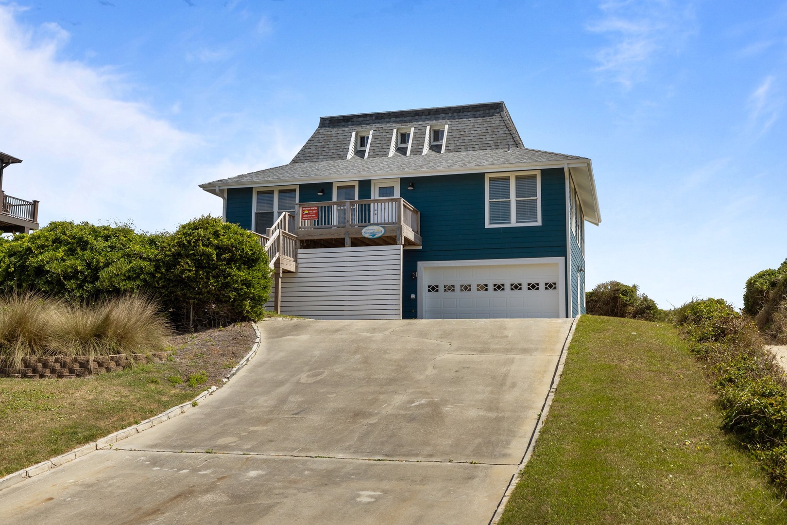 Charming coastal home with distinctive teal siding and spacious driveway, nestled among natural dunes and greenery.