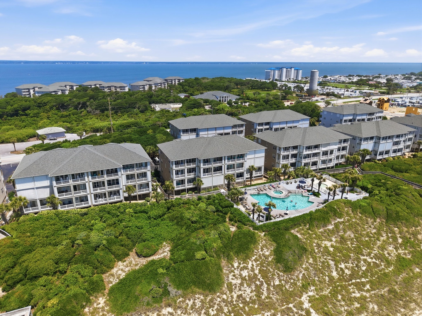 Aerial view of a beachfront vacation rental complex with multiple buildings, a resort-style pool, and lush tropical landscaping situated between the ocean and surrounding coastal neighborhood.