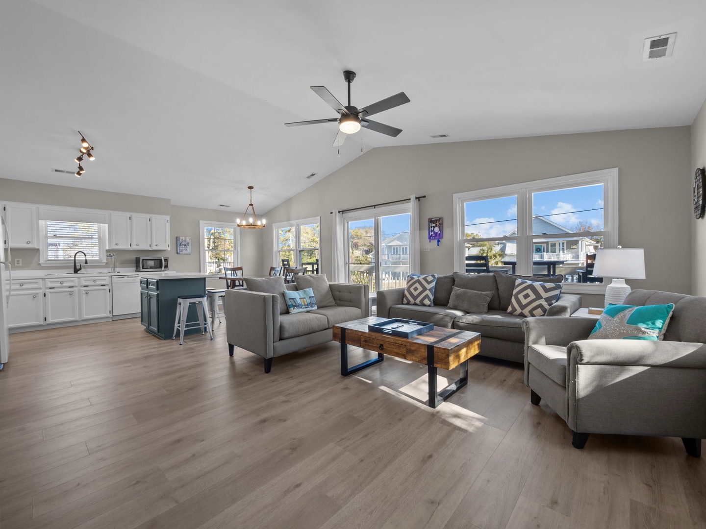 Relax in your spacious open-concept living space featuring modern gray sectional seating, natural light streaming through large windows, and a well-equipped kitchen perfect for preparing meals.