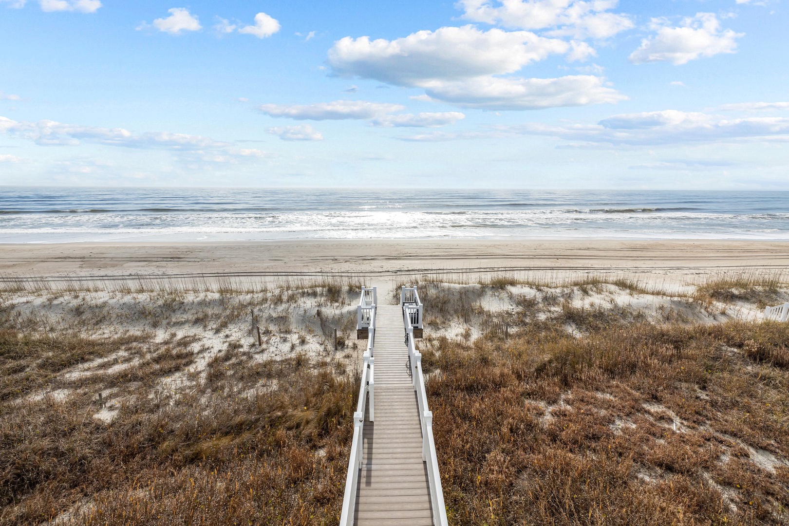 Wooden boardwalk leads through coastal dunes to pristine sandy beach and rolling ocean waves under expansive blue sky.