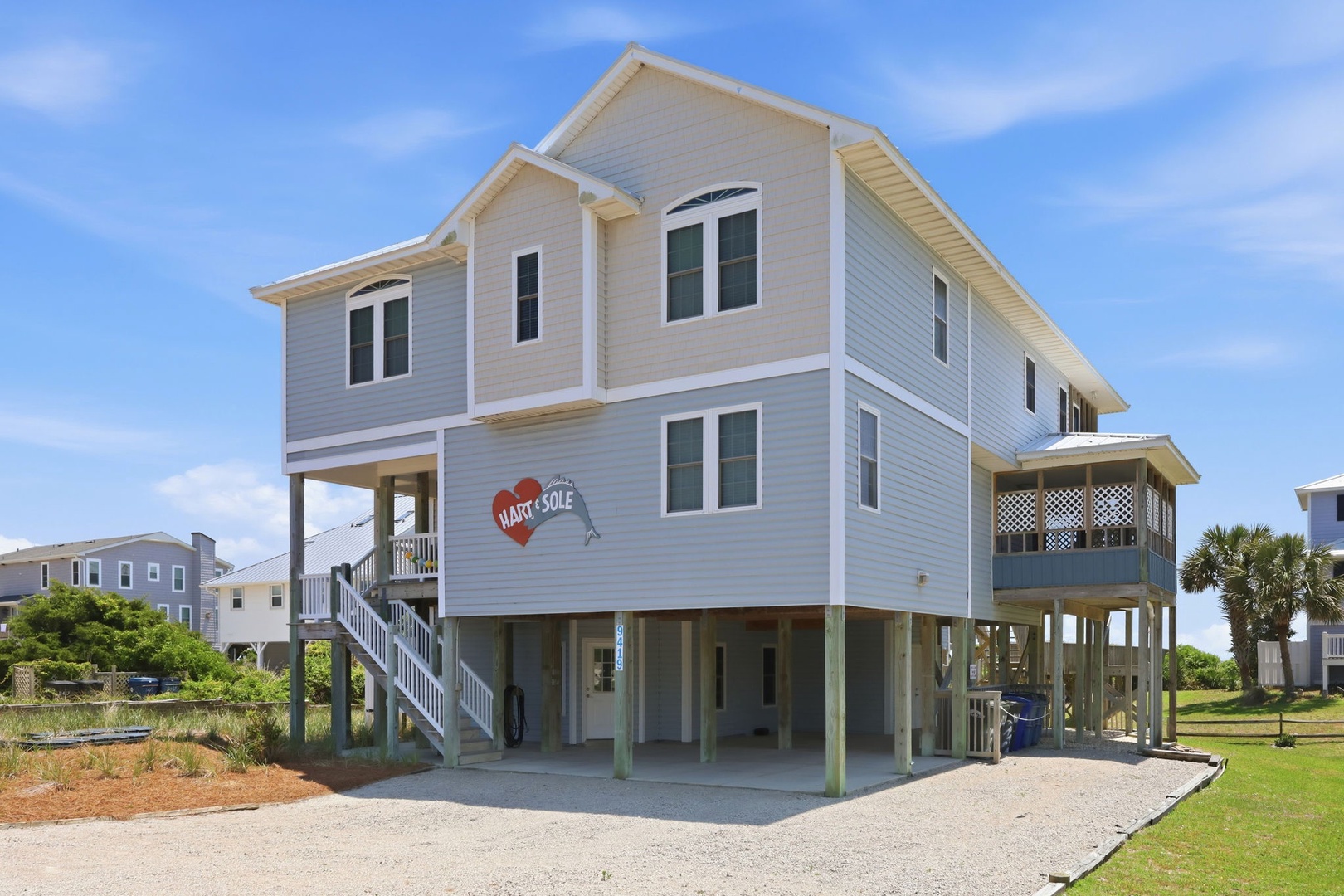Raised coastal home with covered parking and ground-level storage, situated in a quiet residential neighborhood surrounded by palms and greenery.