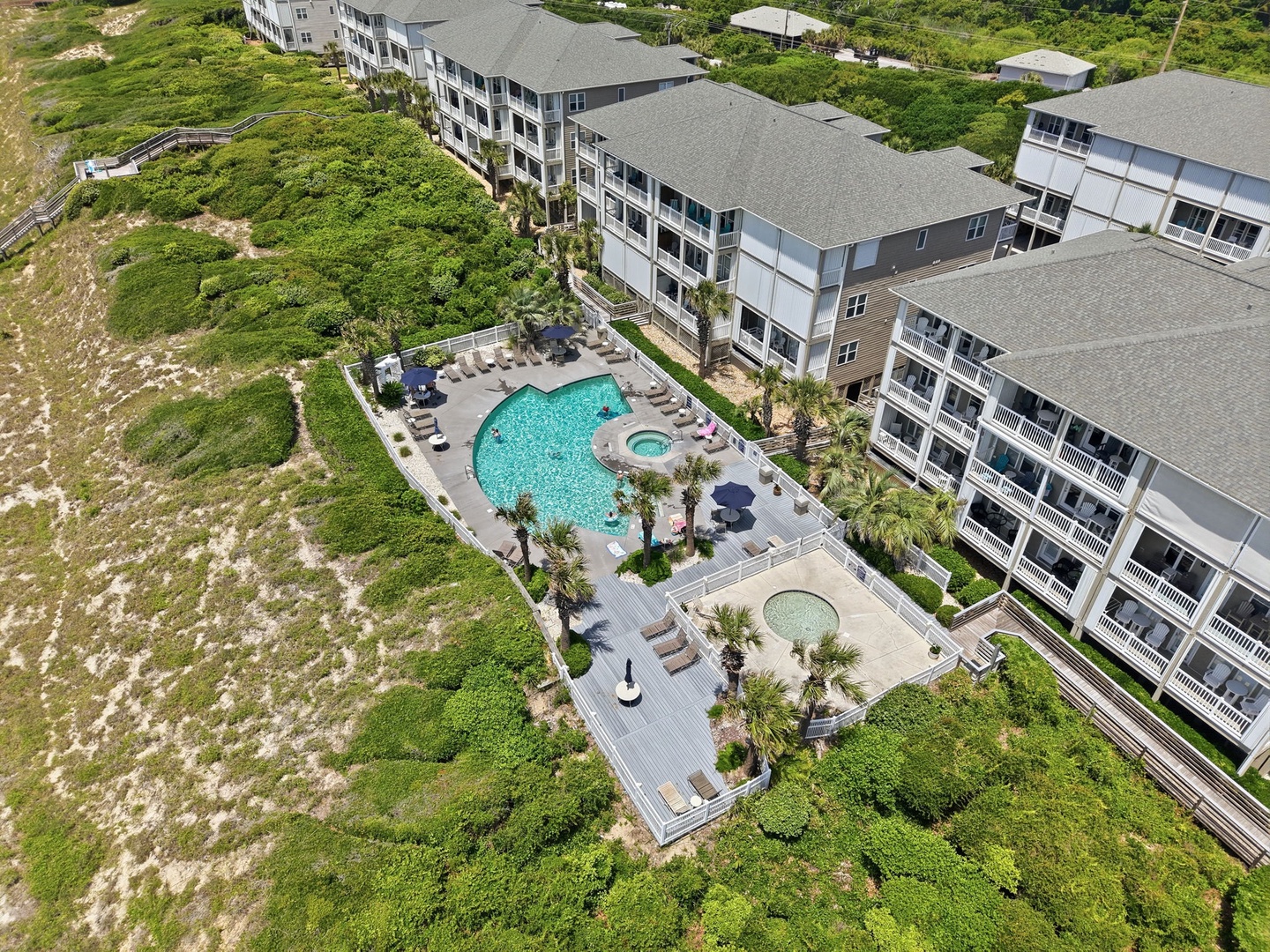 Aerial view showcasing a modern coastal property with two pools, expansive sun decks, and lush landscaping surrounded by natural dunes and greenery.
