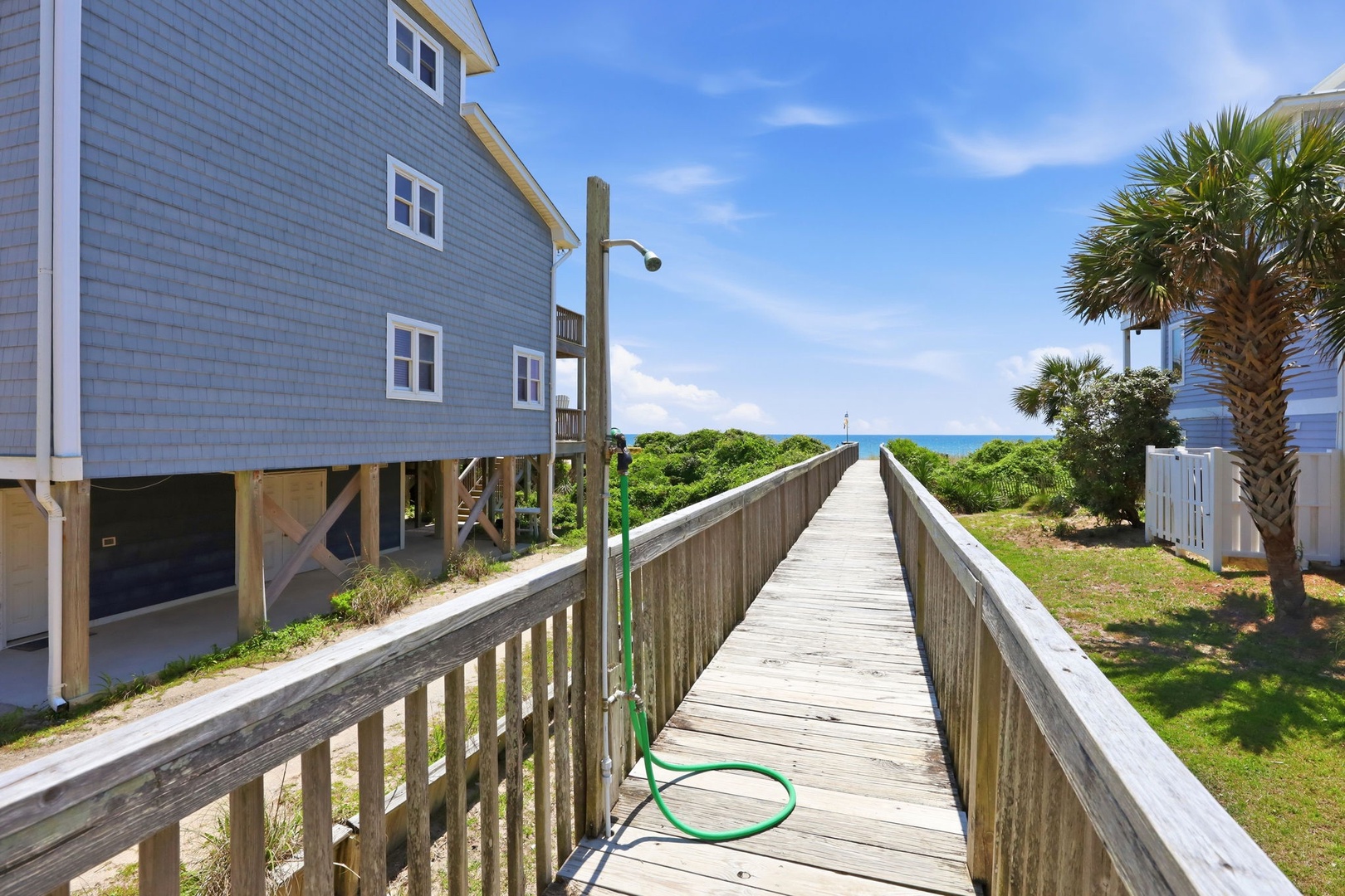 A private boardwalk connects the property directly to the beach, offering convenient coastal access through natural seaside vegetation and past neighboring beach homes with ocean views ahead.