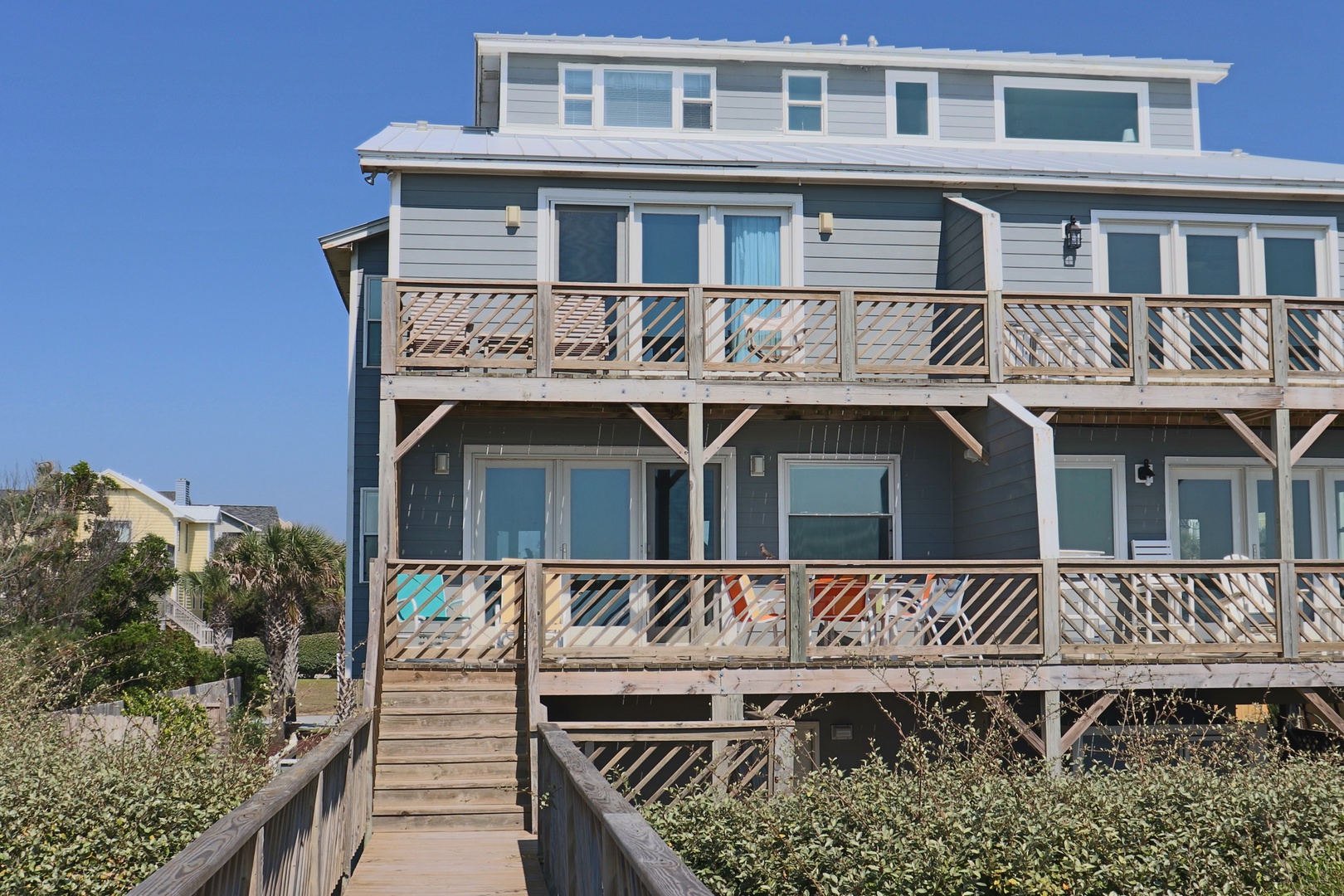 Three-story coastal property featuring multiple decks with decorative railing patterns and elevated boardwalk access through surrounding dune vegetation.