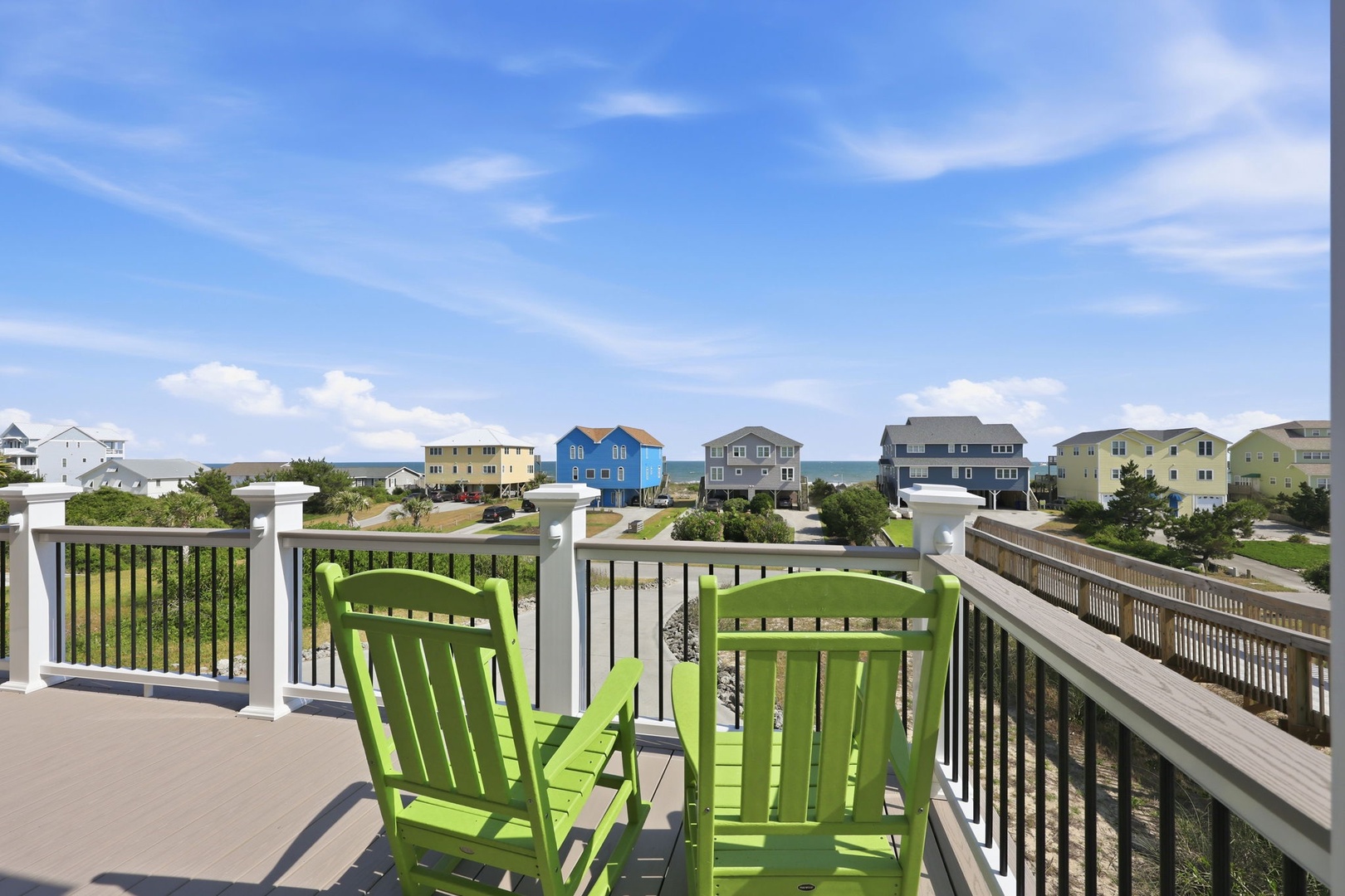 Elevated deck area featuring bright green Adirondack chairs overlooking a residential beachfront community with colorful homes and ocean views beyond.