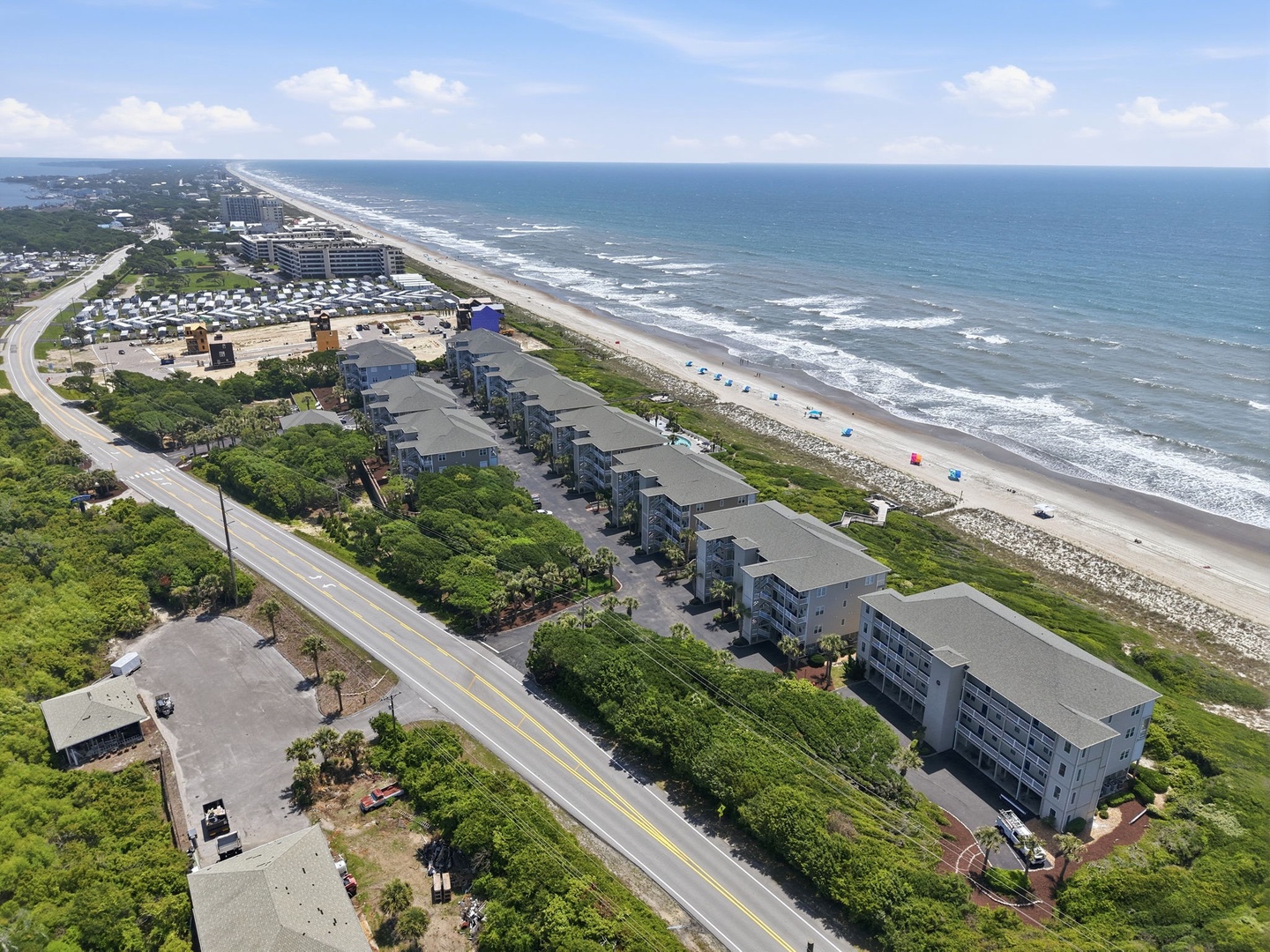 Aerial view of beachfront vacation rental buildings situated along a coastal road with direct beach access, sandy shoreline, and ocean waves visible in the background.
