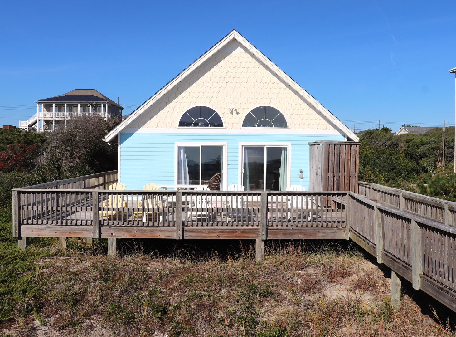 Charming coastal cottage features bright blue siding and spacious wooden deck with outdoor seating, nestled among natural dunes.