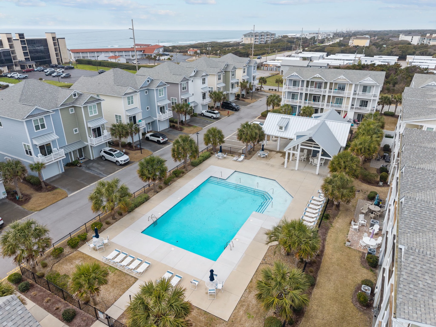 Aerial view of a coastal vacation rental community featuring colorful beach houses, shared swimming pool, and palm-lined streets near the ocean.