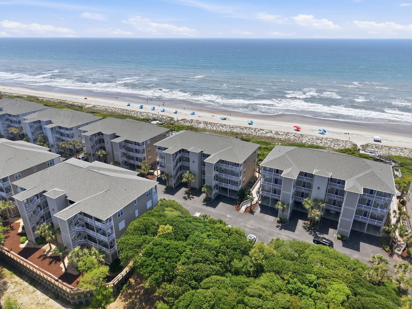 An aerial view of the beachfront property complex situated directly on the sandy shore, featuring multi-story residential buildings with convenient parking and lush landscaping adjacent to the ocean.