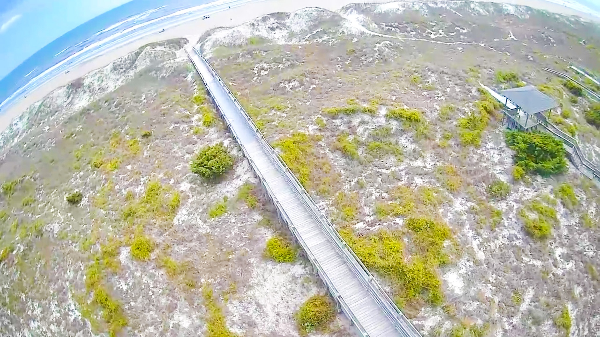 Aerial view reveals a stunning coastal landscape with wooden boardwalk winding through natural dune terrain toward pristine shoreline.