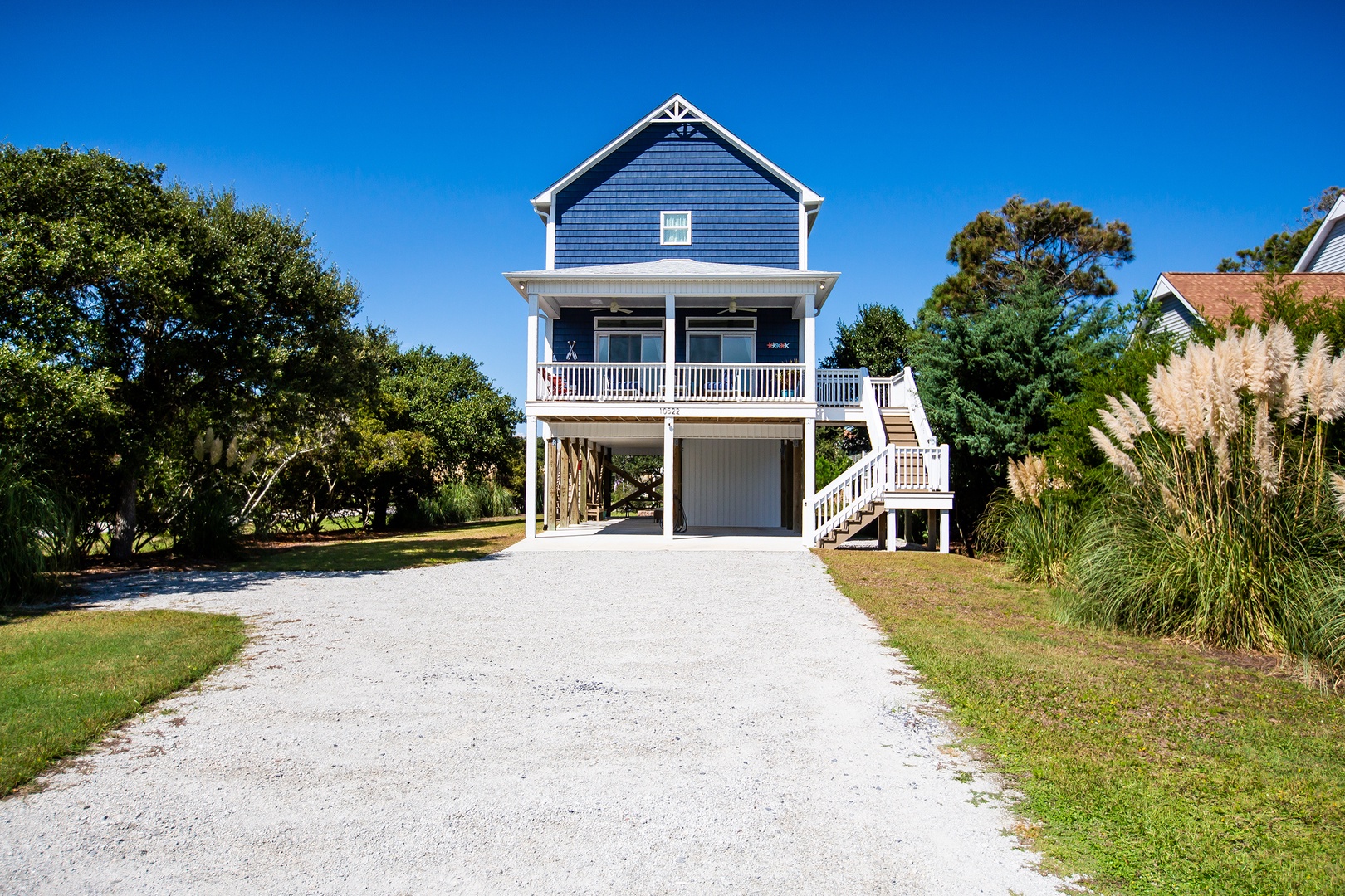 Charming coastal cottage with elevated design and covered parking beneath, surrounded by lush landscaping and mature trees.