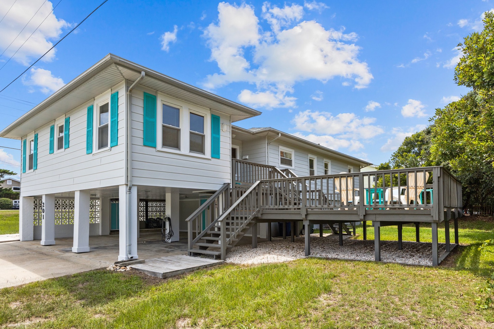 Charming coastal home with spacious deck and parking beneath, surrounded by mature trees under blue skies.