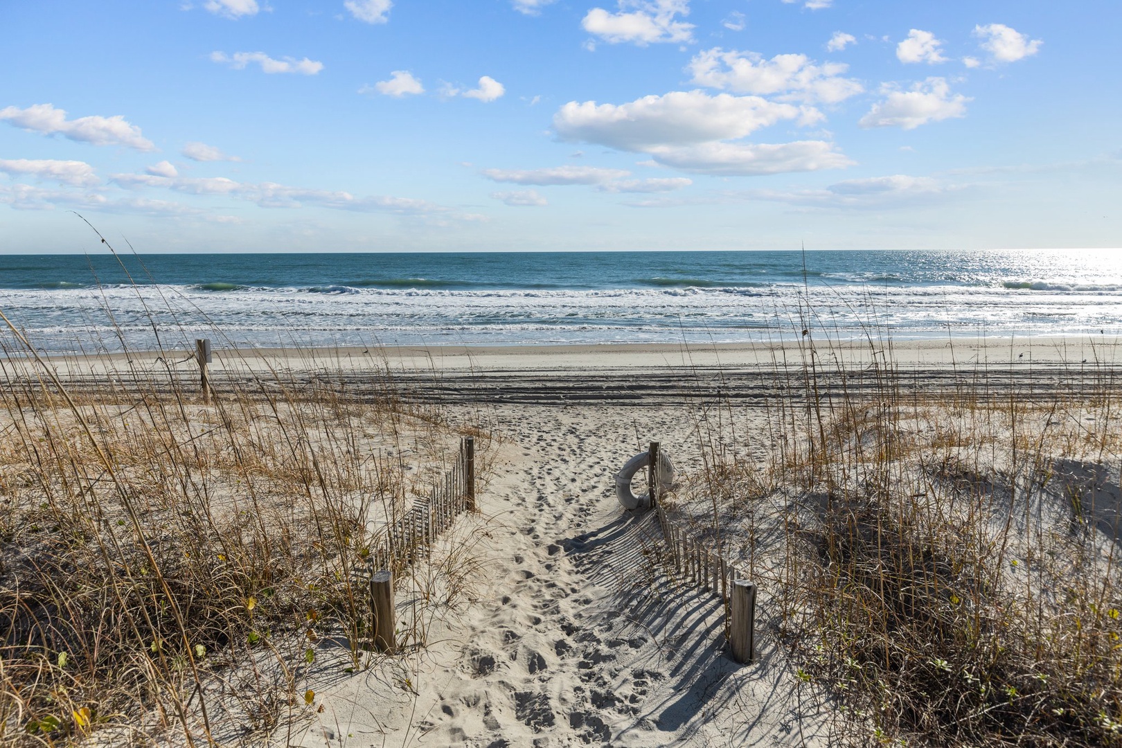 Pristine beach with sandy pathway leading to ocean waves under bright blue skies with scattered white clouds.
