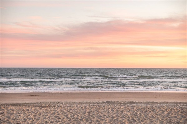 Peaceful beach stretches along the shoreline as soft pink and orange hues paint the evening sky over gentle ocean waves.