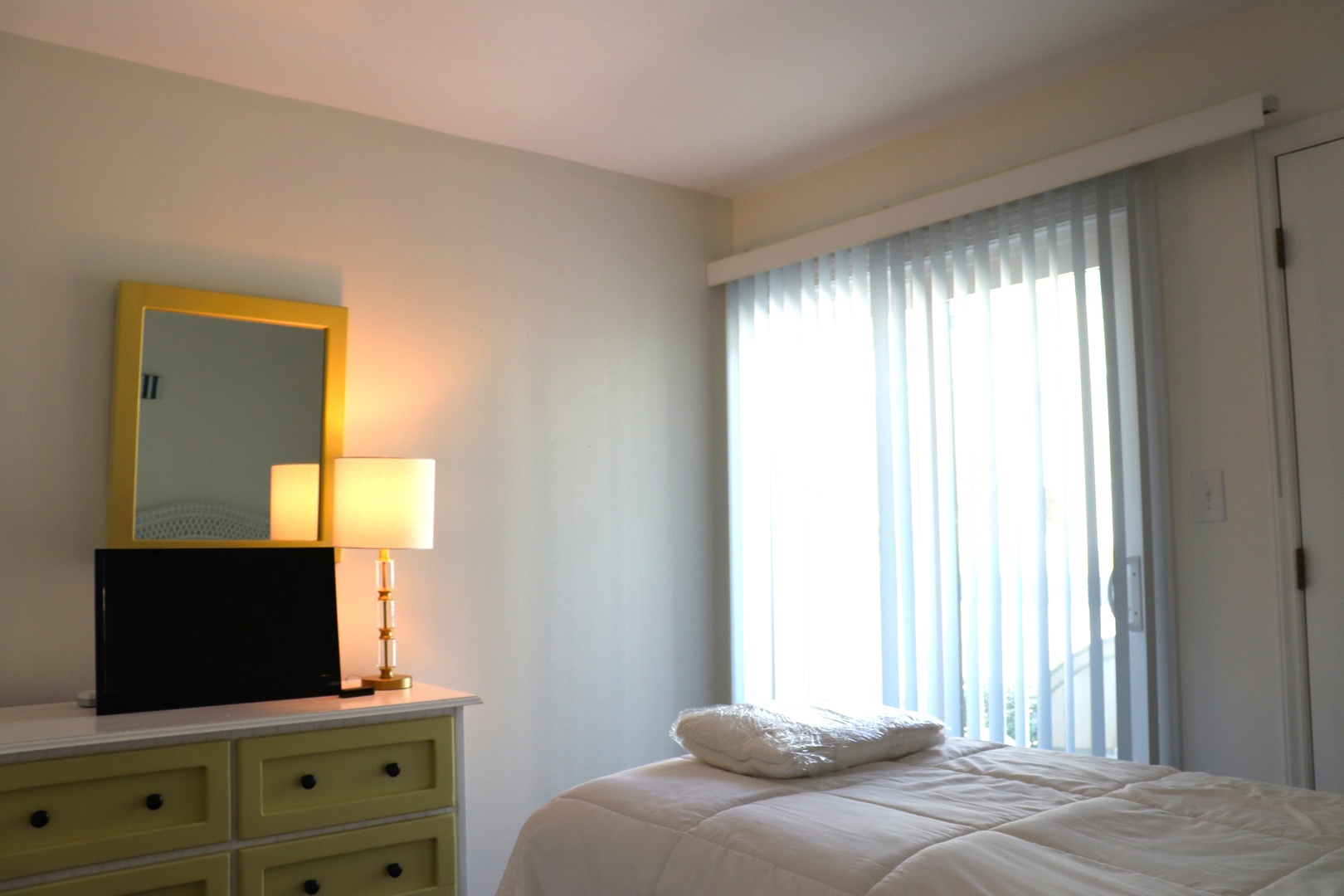 Your peaceful bedroom awaits with crisp linens, warm lighting, and natural light streaming through vertical blinds for the perfect rest.