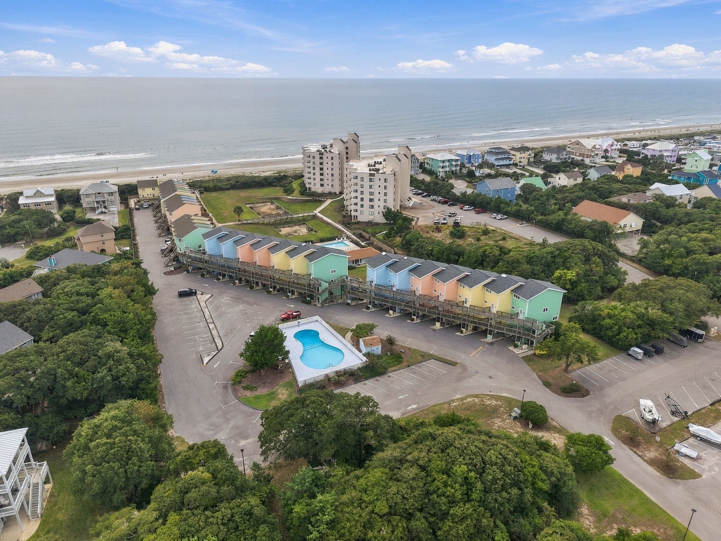 Aerial view of colorful beachfront townhomes nestled among coastal greenery with a community pool, just steps from the sandy shore and nearby high-rise towers.