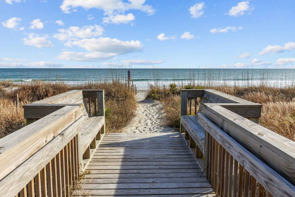 Wooden boardwalk leads through coastal dunes to pristine beach access with ocean views and sandy pathways.