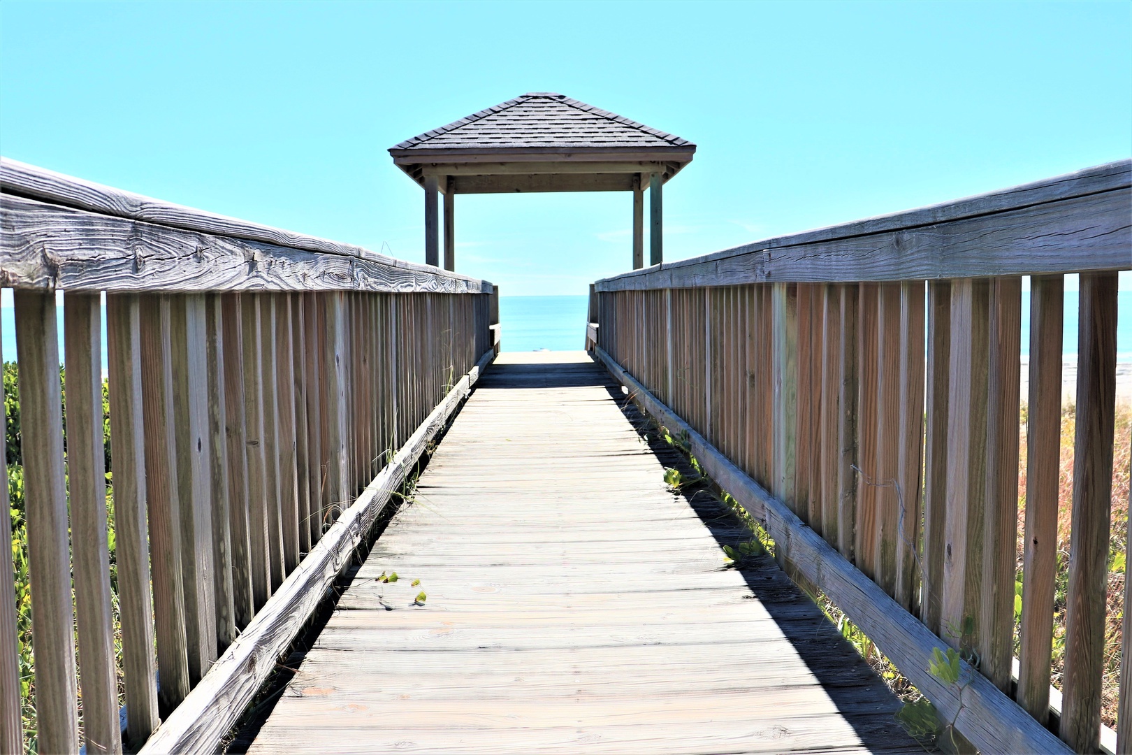 A wooden boardwalk extends toward a covered pavilion and the ocean beyond, offering direct beach access from the property.