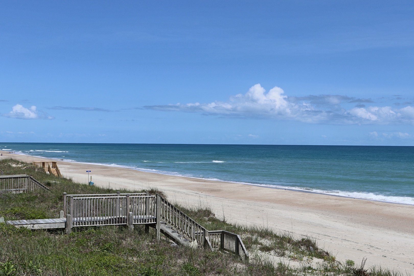 Pristine sandy coastline stretches endlessly with gentle waves and crystal-clear turquoise waters under expansive blue skies dotted with fluffy white clouds.