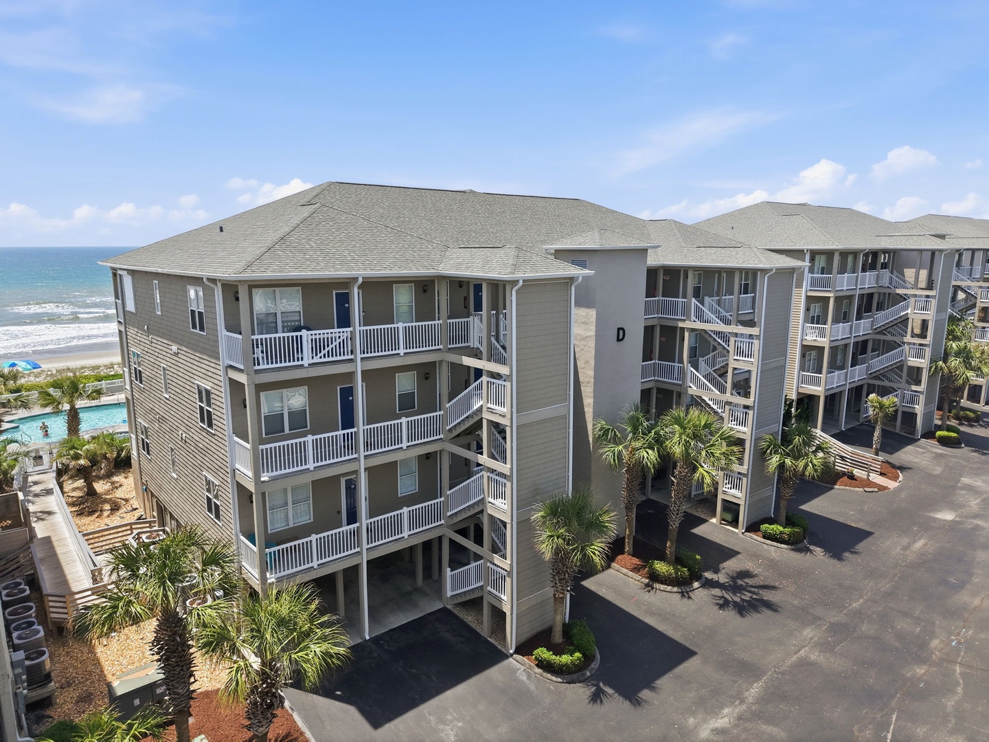 Oceanfront vacation rental complex with covered parking, private balconies, and direct beach access framed by tropical palms and nearby pool facilities.