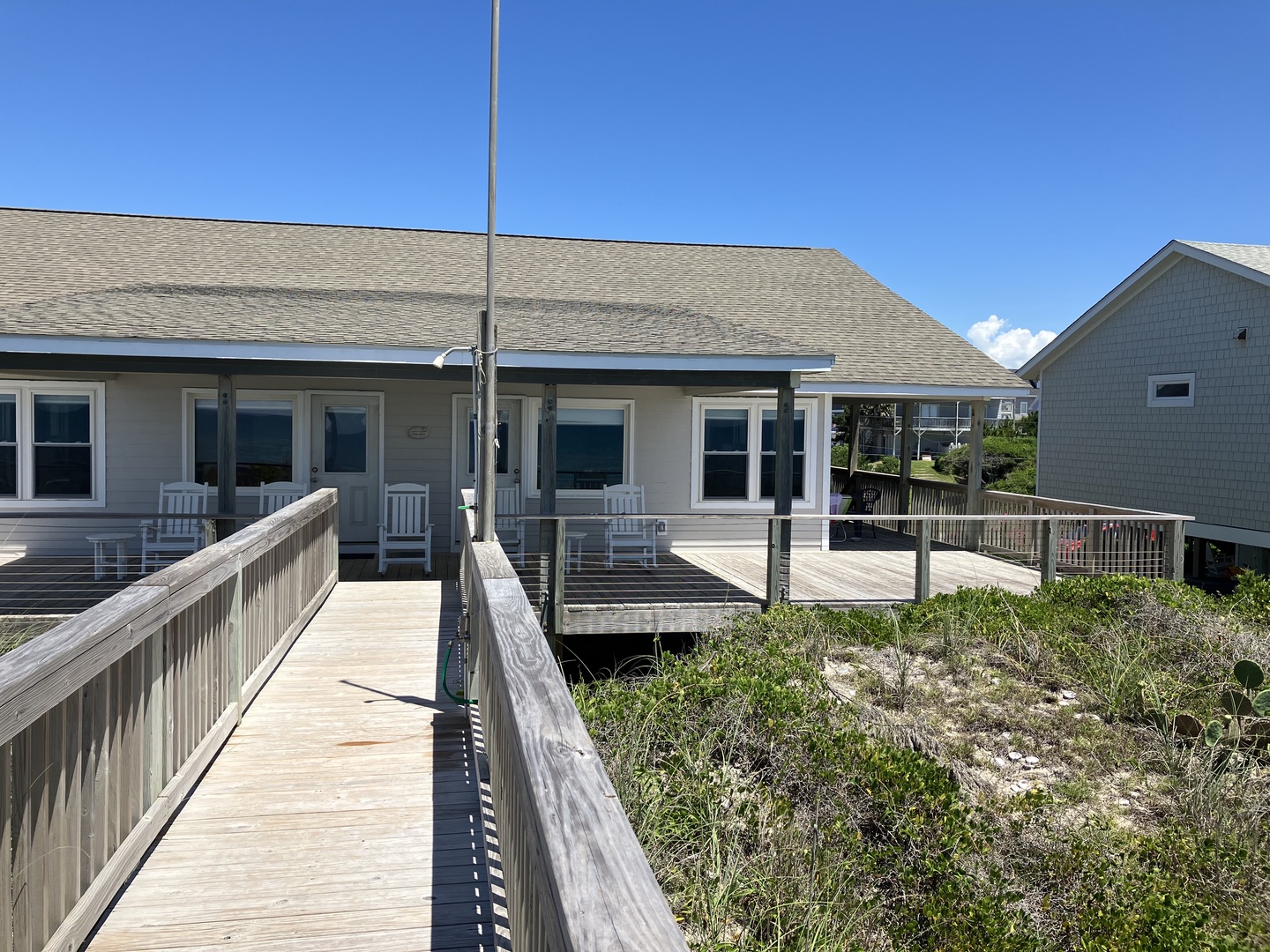 Charming beachside cottage with elevated deck and coastal architecture, surrounded by natural dune vegetation under clear blue skies.