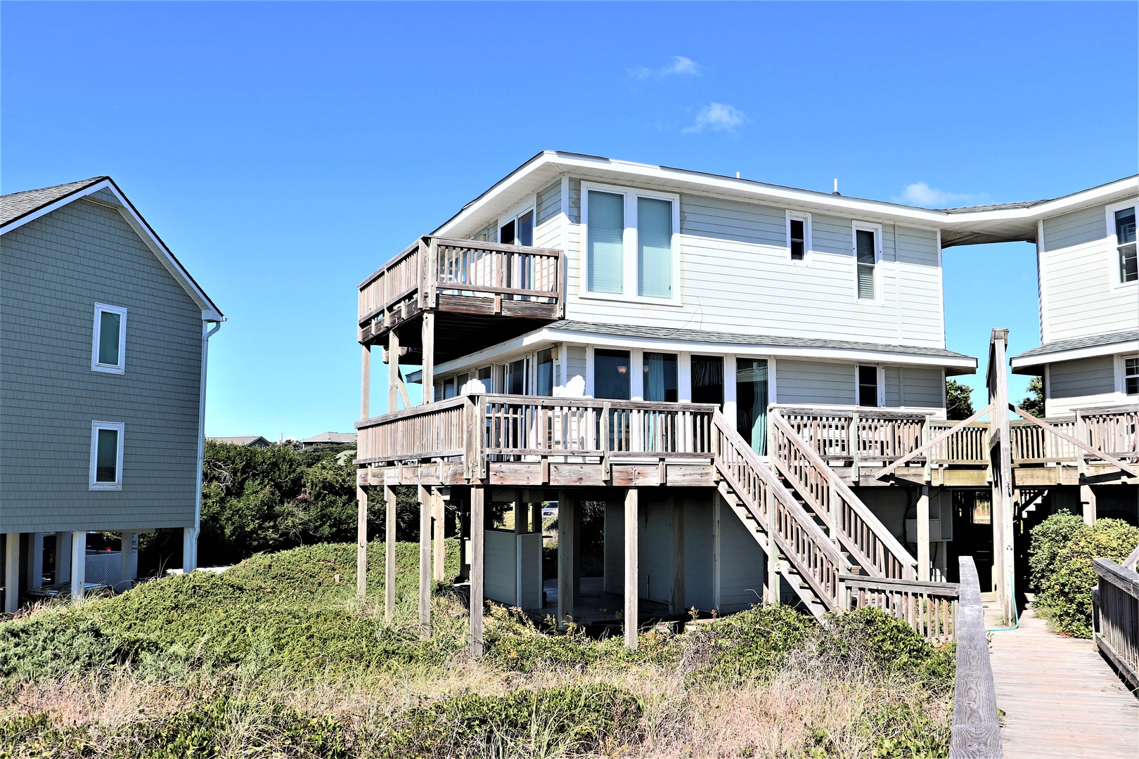 Beach house with elevated wooden decks and walkways surrounded by coastal vegetation under clear blue skies.