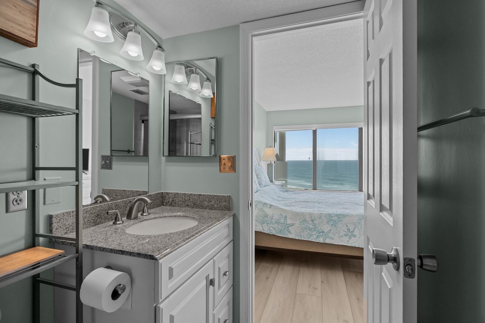Your en-suite bathroom features granite countertops and modern vanity, with glimpses of ocean views from your cozy bedroom beyond.
