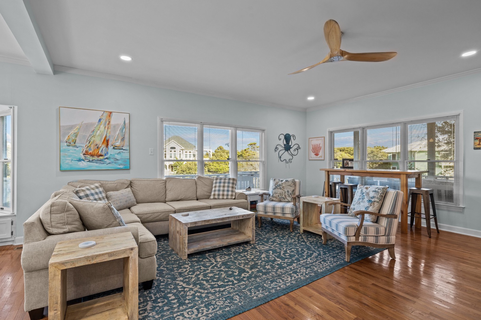 Relax in your bright, coastal-themed living room with plush seating, nautical artwork, and panoramic windows showcasing beautiful neighborhood views.