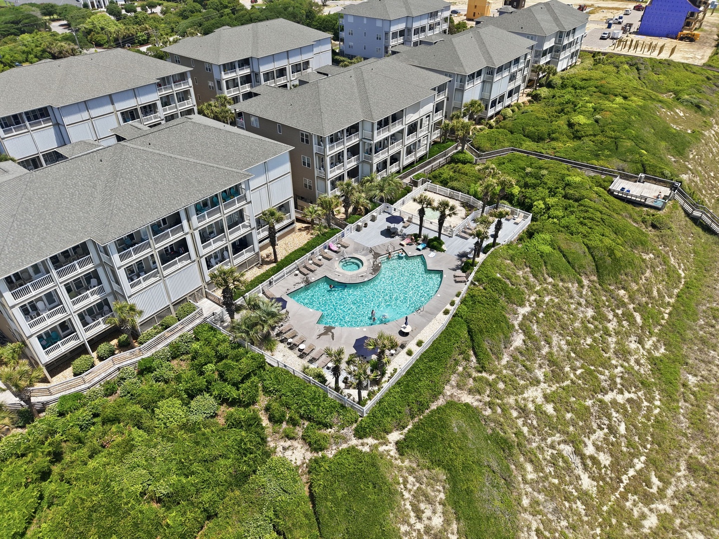 Aerial view of the vacation rental property featuring multi-story buildings surrounding a central pool area with hot tub, bordered by natural coastal landscape and walkways.
