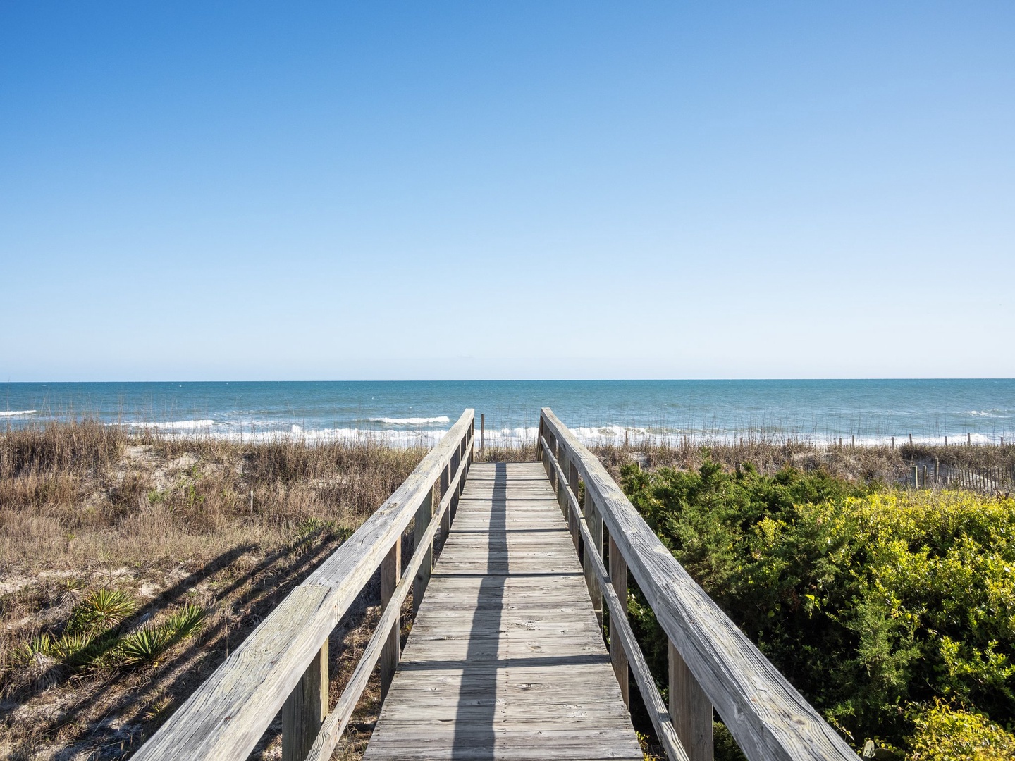 Wooden boardwalk leads through coastal dunes to pristine beach with turquoise waters and clear blue skies.