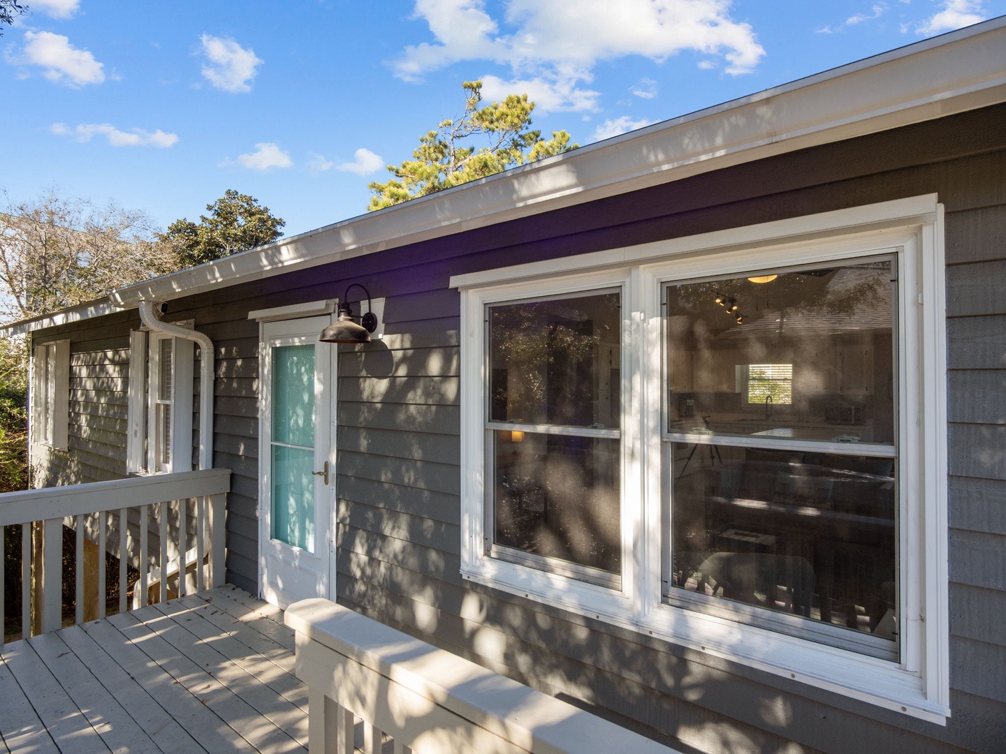 Charming cottage exterior features gray shingle siding, bright turquoise door, and wraparound deck surrounded by mature trees.