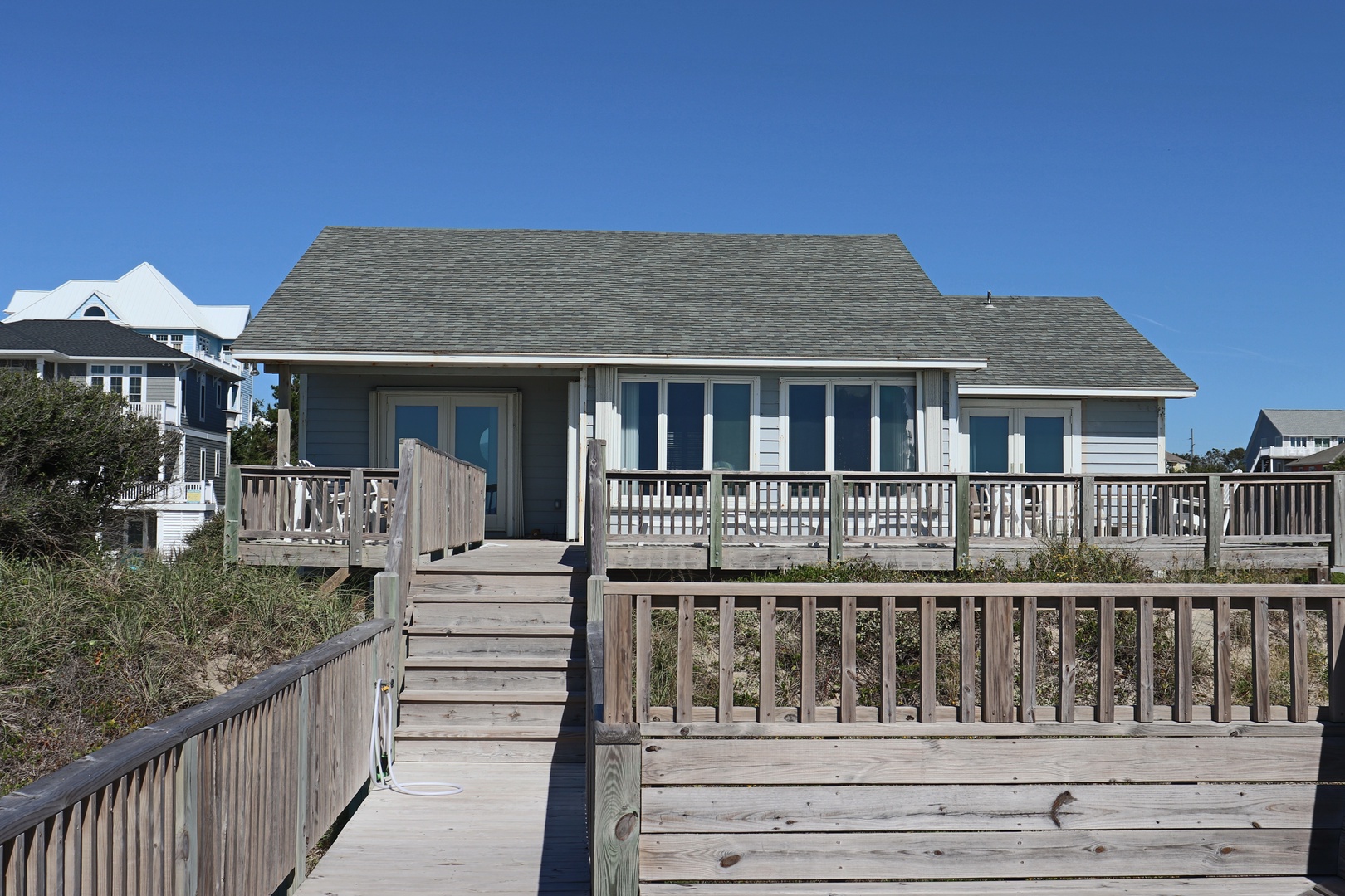 Charming beachfront cottage features wraparound deck with ocean access walkway, perfect for coastal getaways under clear blue skies.
