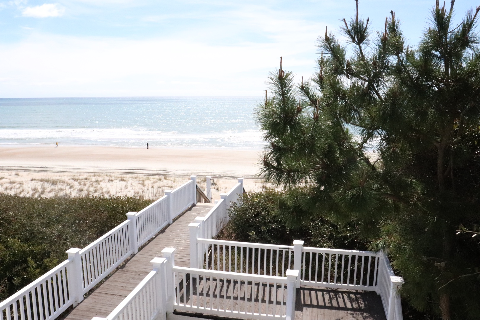 Wooden boardwalk leads through coastal dunes to pristine sandy beach with gentle ocean waves and expansive horizon views.