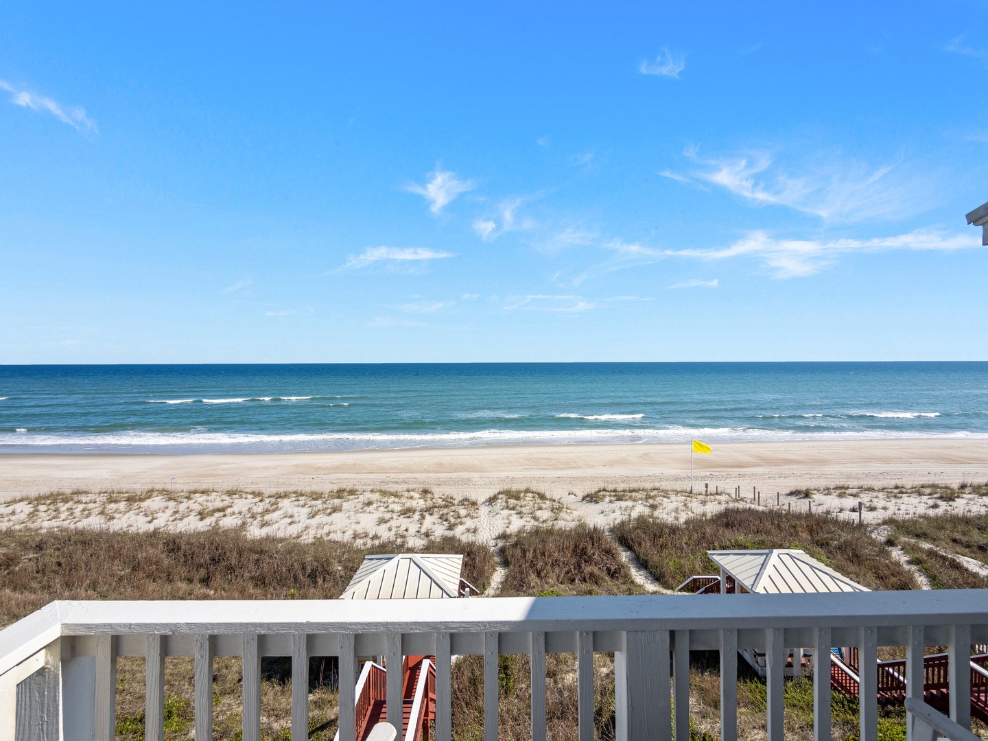 Spectacular oceanfront view showcases pristine sandy beach with gentle waves rolling to shore beneath expansive blue sky dotted with wispy clouds.