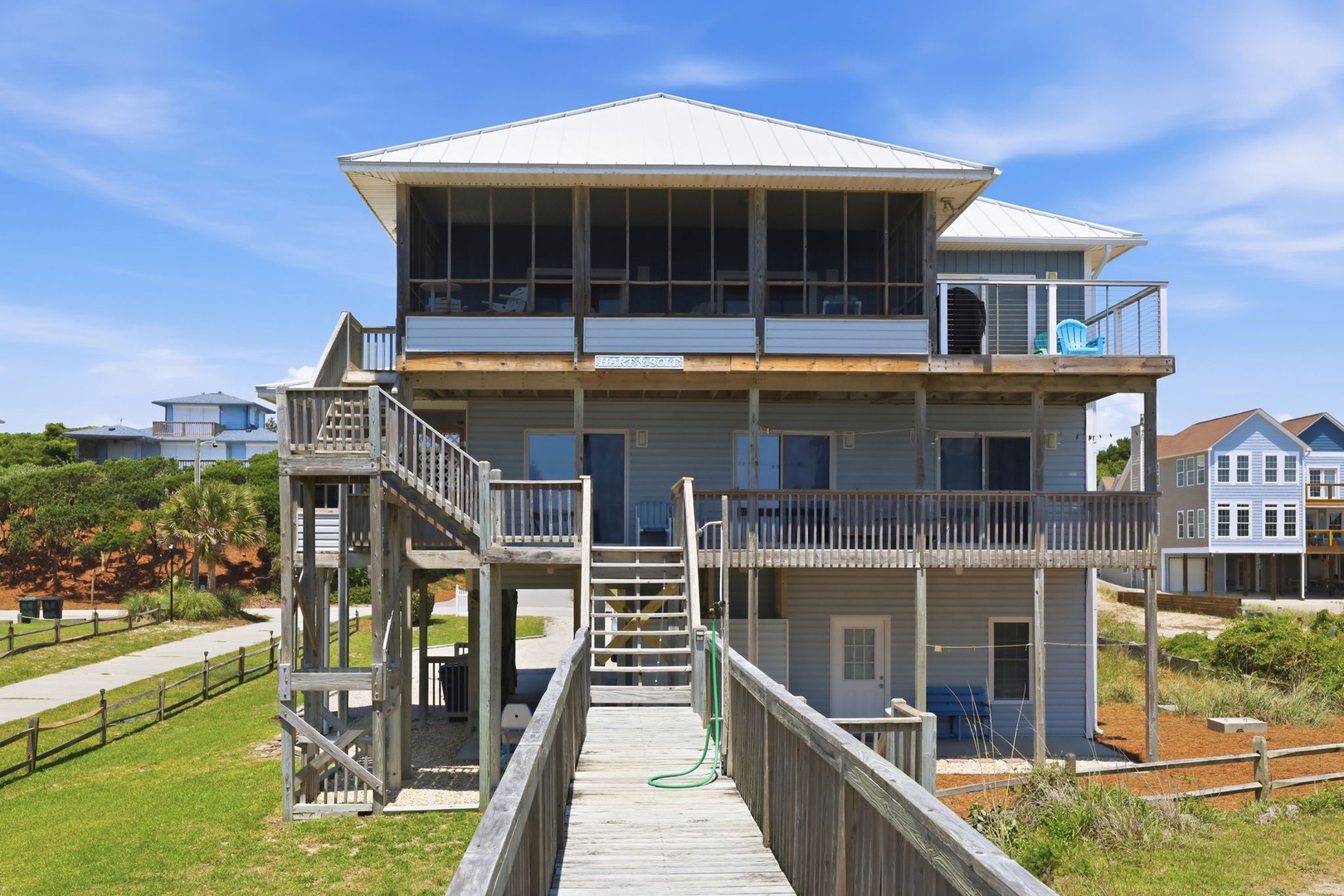 Elevated coastal beach house with multiple levels, screened porch, expansive deck spaces, and weathered wood boardwalk leading to the entry—ideal for a seaside vacation retreat.