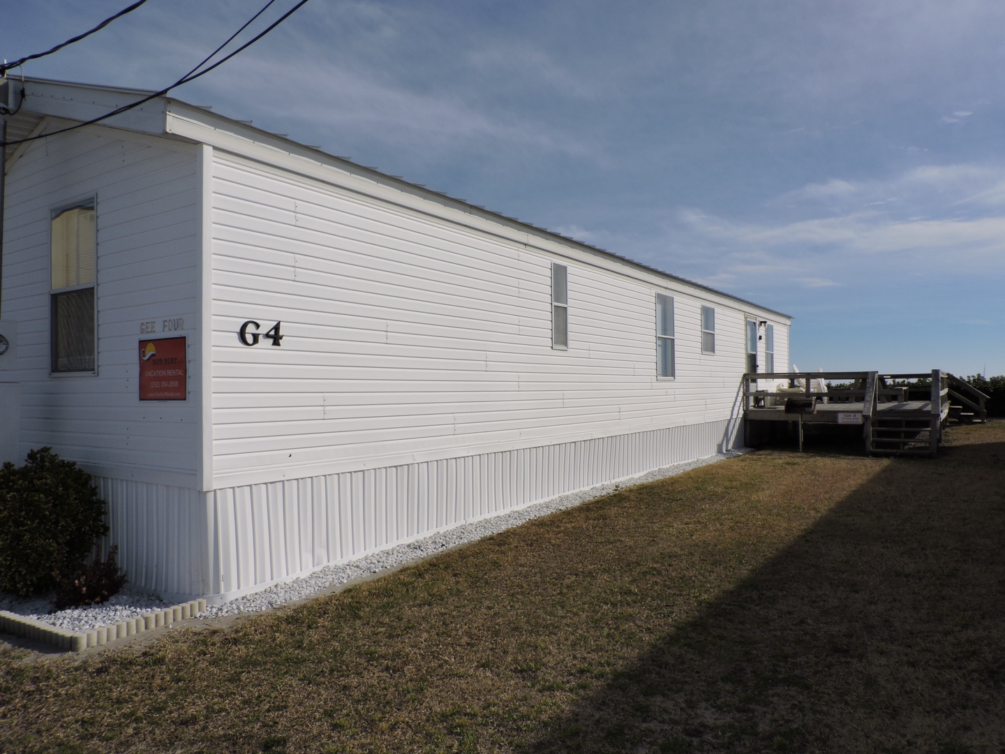White mobile home unit 64 with wooden deck entrance, situated in peaceful residential setting with maintained grounds.