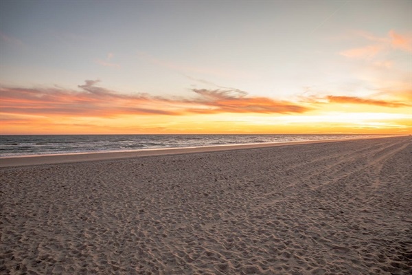 Golden sunset paints the sky over pristine sandy beach with gentle waves rolling to shore.